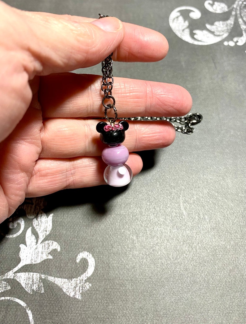 May include: A black and purple beaded necklace with a black and pink Minnie Mouse charm. The necklace is on a black chain.
