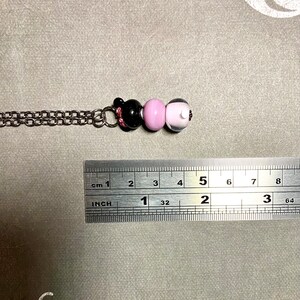 May include: A silver chain necklace with three glass beads. The beads are black, pink, and white. The necklace is approximately 5 cm long.