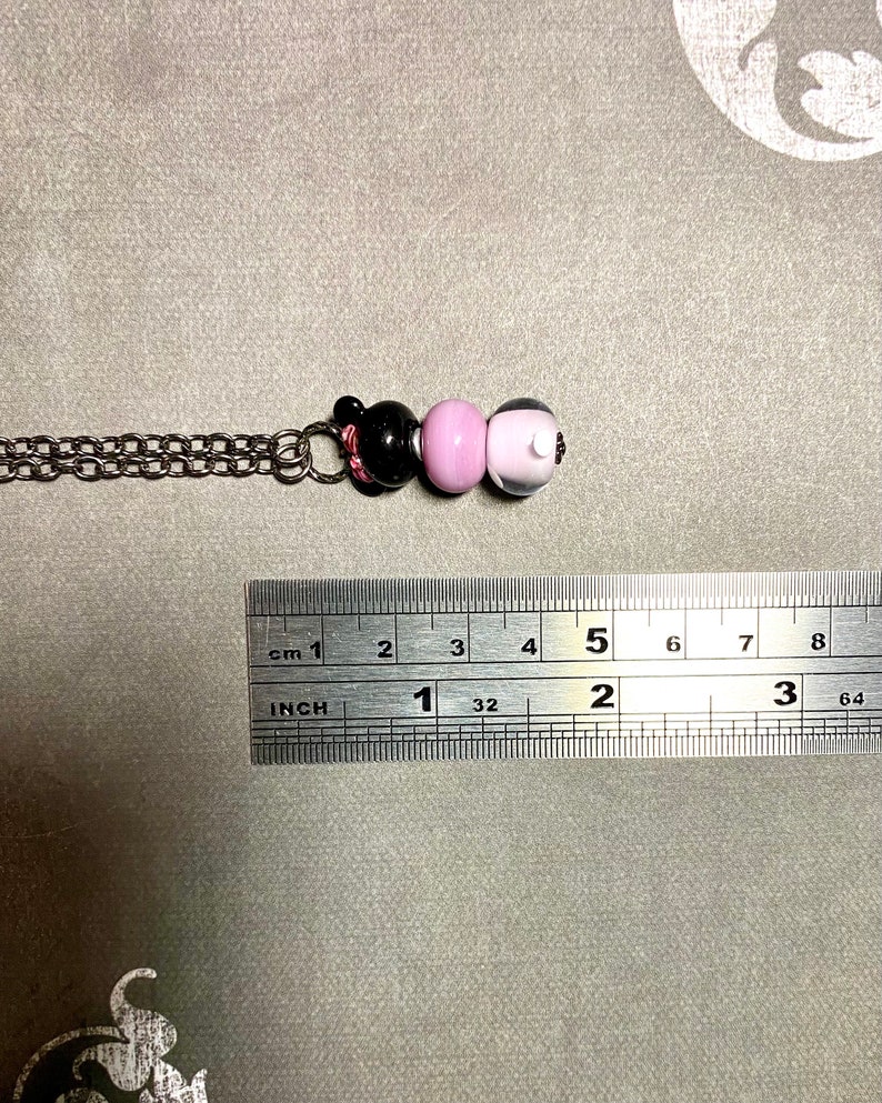 May include: A silver chain necklace with three glass beads. The beads are black, pink, and white. The necklace is approximately 5 cm long.