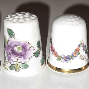 Puede incluir: Cuatro dedales de porcelana blanca con varios diseños florales y frutales. Uno presenta un pájaro azul, otro una flor morada, un tercero una guirnalda floral y el último, un racimo de frutos morados. Cada dedal tiene una parte superior texturizada.