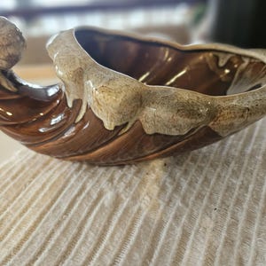 Puede incluir: Un cuenco de cerámica con forma de concha marina con un esmalte marrón y beige. El cuenco tiene un diseño curvo y abierto con una superficie texturizada. El borde y el asa son beige, mientras que el interior y el cuerpo son marrones.