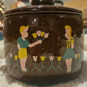 Puede incluir: Una fuente de cerámica marrón con tapa. El plato está decorado con una escena pintada de dos figuras, una de ellas sosteniendo flores. Flores amarillas y rosas están pintadas alrededor de la base del plato.