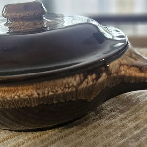 Puede incluir: Una cazuela de cerámica marrón con tapa y asa. El plato tiene un acabado brillante con una parte superior marrón oscuro y una banda texturizada marrón claro en el medio. El asa también es marrón y texturizada.