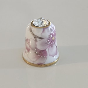 May include: A decorative thimble with a floral design. The thimble is white with pink and purple flowers and gold trim. A clear, faceted gem is set at the top. A sewing accessory.