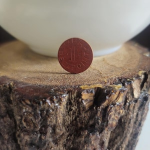 Op de afbeelding: Een kleine, ronde, roodbruine token met het nummer "1" in het midden en de woorden "SPA POINT" rondom. De token ligt op een getextureerd, bruin houten oppervlak, met een witte kom op de achtergrond.