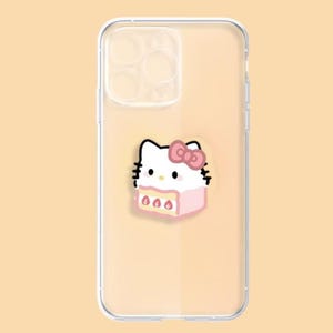 May include: Clear phone case featuring a cartoon illustration of a white cat with a pink bow and a pink cake. The case is designed to fit a smartphone, with cutouts for the camera and buttons. The background is a light peach color.
