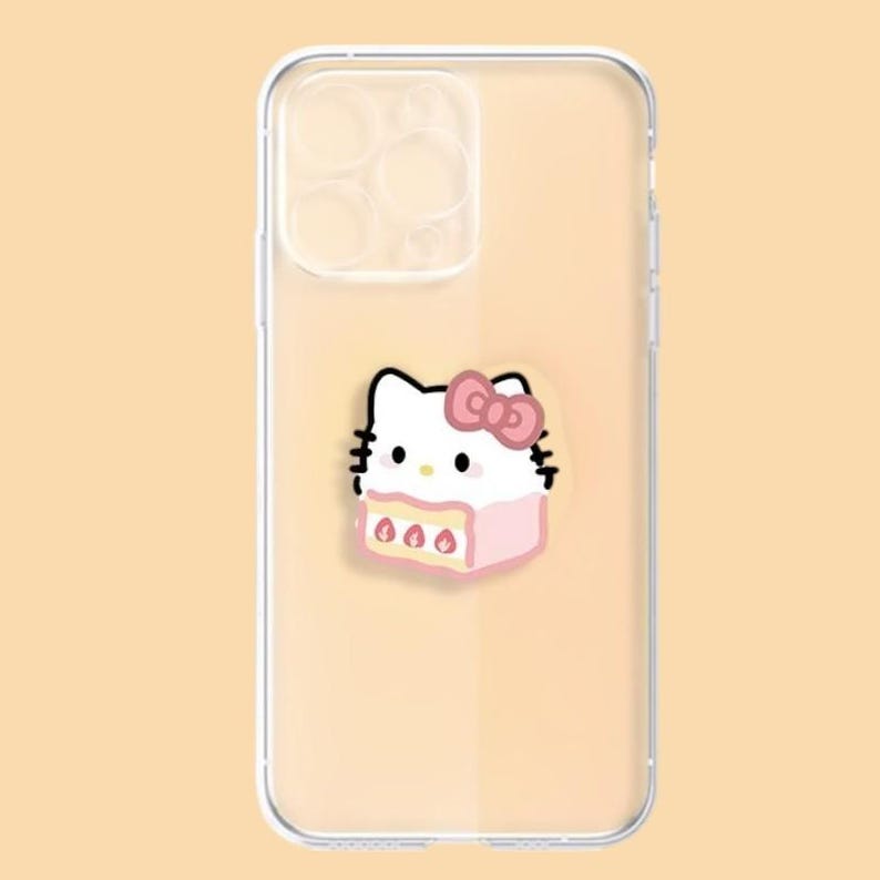May include: Clear phone case featuring a cartoon illustration of a white cat with a pink bow and a pink cake. The case is designed to fit a smartphone, with cutouts for the camera and buttons. The background is a light peach color.