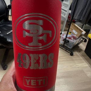 May include: A red Yeti tumbler featuring the San Francisco 49ers logo and the text "49ERS". The stainless steel tumbler has a silver rim and base. The Yeti logo is at the bottom.