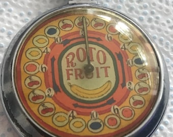 May include: A round silver-toned metal pocket watch-style game with a glass cover. The dial features the text ROTO FRUIT, a yellow banana illustration, and a circular border of fruit icons including cherries, lemons, and plums with numbers.