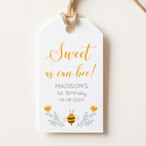 May include: A white gift tag with the text "Sweet as can bee!" in gold script. The tag includes the name "Madison's" and "1st Birthday" with a date. A cartoon bee and floral design are at the bottom. A natural twine string is attached.