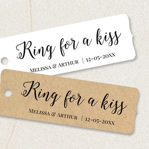 May include: Two rectangular tags with rounded corners. The top tag is white, and the bottom tag is brown. Both tags have the text "Ring for a kiss" in elegant script. Below the text is "MELISSA & ARTHUR | 12-05-20XX".