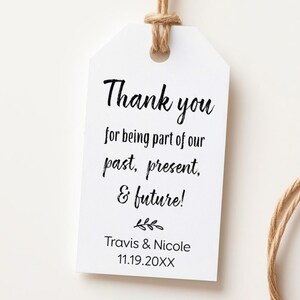 May include: A white rectangular paper gift tag with rounded top corners, featuring black printed text that reads: Thank you for being part of our past, present, & future! Travis & Nicole 11.19.20XX. A small leaf icon sits above the names, and brown twine is attached.