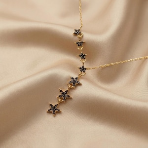 May include: A gold-tone necklace featuring a delicate chain and a vertical drop design adorned with seven small star-shaped charms. Each star is encrusted with tiny black stones, creating a contrast against the metallic gold finish of the jewelry.