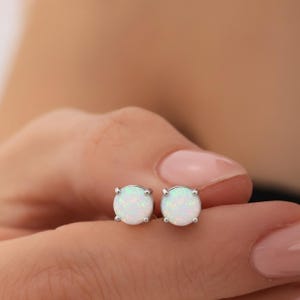 Può includere: Un paio di orecchini a lobo rotondi in opale bianco con montatura in argento. Gli opali hanno una sottile lucentezza iridescente. Gli orecchini sono esposti su una mano.