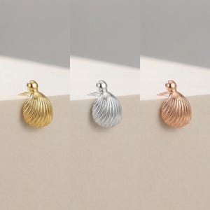 May include: Three pairs of stud earrings in gold, silver, and rose gold. Each earring has a textured, shell-like design with a small post for piercing. The earrings are displayed on a neutral surface, showcasing their different metallic finishes.