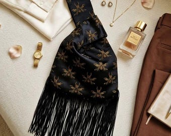 May include: A cream-colored collared shirt, brown trousers, black patent leather pumps, and a black satin handbag with gold embroidered beetle motifs and long black fringe. Accessories include a gold watch, gold earrings, a gold necklace, and perfume.
