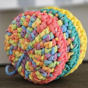 May include: A colorful crocheted ball with a rainbow pattern. The ball is made of yarn in shades of pink, yellow, green, blue, and purple.