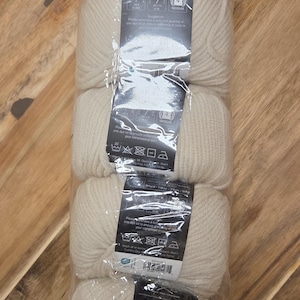 May include: A package of off-white Essential Wool yarn skeins. Each skein is 1.8 oz (50g) and 84 yards (77m). The packaging includes care instructions and fiber content information.