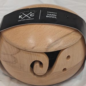 May include: A light-colored wooden yarn bowl with a carved swirl design and two small holes. The bowl has a black band around it with the text "KXC KNIT AND CROCHET" and "YARN BOWL".