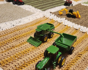 May include: A green toy tractor and a green toy combine harvester on a brown and yellow crocheted rug. The rug is divided into squares, each with a different color and texture.