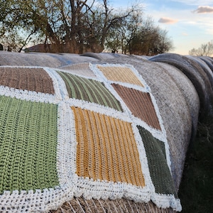 May include: A crocheted blanket with a patchwork design in shades of green, brown, and yellow. The blanket is laid out on a stack of hay bales.