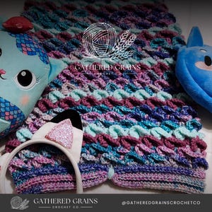 Op de afbeelding: Een gehaakt deken in tinten van paars, blauw en turquoise. Het deken is gemaakt met een textuur-steek en heeft een "Gathered Grains Crochet Co." logo in het midden.