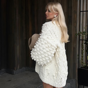 May include: A woman wearing a cream-coloured, chunky knit cardigan with large, textured sleeves. She is holding a beige hat. The background features a wooden door and wall.