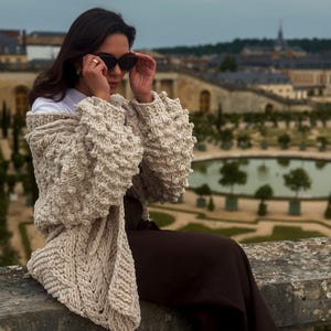 May include: A beige, chunky knit cardigan with textured sleeves and a patterned front. The cardigan is draped over a person wearing sunglasses and a white shirt. The background features a scenic view of a garden and buildings.