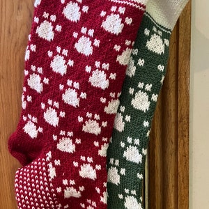 May include: Two knitted Christmas stockings with a paw print design. One stocking is red with white paw prints and the other is green with white paw prints.