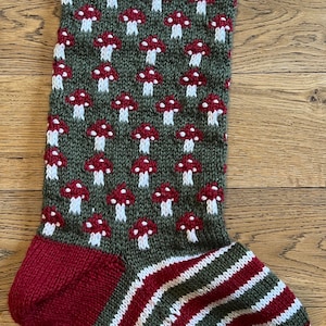May include: Hand-knitted Christmas stocking with a white top and a burgundy cuff. The body features a pattern of red and white mushrooms on a green background, with burgundy, white, and green stripes on the foot. A festive holiday decoration.