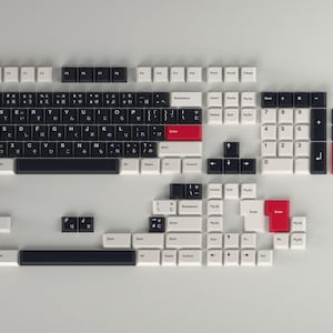 以下が含まれることがあります： 白、黒、赤のキーキャップが付いた分解されたメカニカルキーボード。キーは白い表面に配置され、キーボードの個々のコンポーネントを示しています。レイアウトには、英数字キー、テンキー、ファンクションキーが含まれています。
