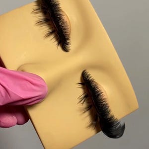 May include: A tan-colored practice face with closed eyes and long, black eyelashes. The eyelashes are thick and full, with a curved shape. The face is held by a person wearing a pink glove.