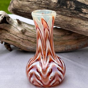 Puede incluir: Un pequeño jarrón decorativo de vidrio con base bulbosa y cuello estrecho. El jarrón presenta un patrón arremolinado de colores blanco y marrón rojizo. El jarrón mide aproximadamente 13 cm de altura, perfecto para exhibir flores pequeñas o como acento decorativo.