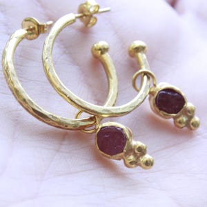 May include: A pair of gold hoop earrings with a hammered texture, each featuring a dangling charm composed of a single deep red, irregular gemstone set in gold, accented by three small gold spheres at the base of the stone setting.