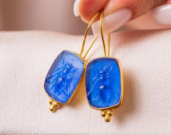May include: A pair of gold earrings with blue glass bee charms. The earrings are square shaped and have a delicate gold chain.