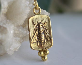 May include: A rectangular gold-toned pendant featuring a raised relief of a bee in the center. The pendant has a small circular bail at the top and three small spherical beads attached to the bottom edge, creating a textured metallic finish.