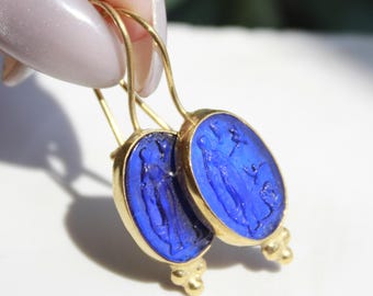 May include: A pair of gold-tone earrings with oval, cobalt blue insets. Each inset features a carved figure. The earrings have a hook closure and a decorative, three-ball design at the bottom.