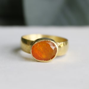 May include: A gold ring with an oval, orange gemstone. The ring has a wide band and a bezel setting that holds the gemstone securely. The gemstone has a faceted surface that catches the light, creating a sparkling effect.