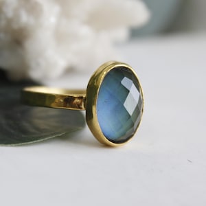 Op de afbeelding: Een gouden ring met een ovale, facetgeslepen, blauwgroene edelsteen. De ring heeft een gehamerde textuur op de band en een gladde, gepolijste bezel setting. De edelsteen heeft een kleurengradiënt van lichtblauw tot diepgroen.
