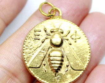 May include: A gold-colored round pendant featuring a detailed bee design. The pendant has a loop at the top for hanging and includes embossed symbols and letters around the bee. The bee's wings and body are clearly defined.