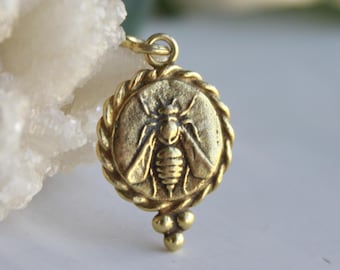 May include: A gold-colored pendant with a detailed bee design. The round pendant has a braided border and a small cluster of three beads at the bottom. The bee is centered within the pendant, showcasing its wings and body.