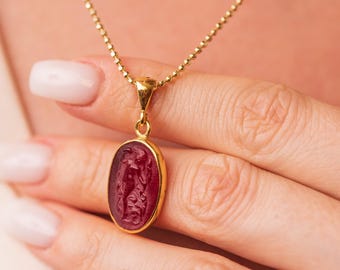May include: A gold-colored necklace with a pendant featuring a red oval stone. The stone has a carved design. The necklace is worn around the neck.