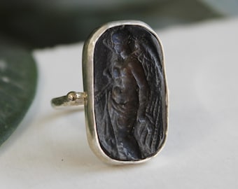 May include: Silver ring featuring a dark, rectangular stone with an engraved figure. The figure is a classical depiction of a person with flowing garments. The ring has a simple band and a small, rounded setting.