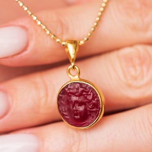 Puede incluir: Collar con colgante dorado con un colgante redondo de piedra roja. El colgante tiene un diseño de rostro tallado y está engastado en un bisel dorado. El collar cuelga de una fina cadena dorada. El fondo está borroso.