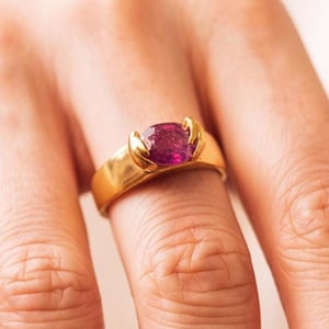 May include: A gold ring featuring a large, oval-cut, deep pink gemstone. The ring's band is a simple, polished gold, with the gemstone held in place by two gold prongs on each side. The ring is worn on a finger.