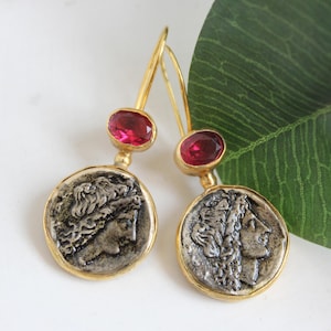 May include: A pair of gold-colored dangle earrings. Each earring features a silver-colored coin with a profile image, a red gemstone, and a gold-colored hook. The earrings are displayed on a white surface with a green leaf in the background.