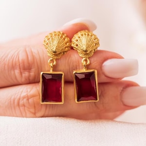 May include: A pair of gold-colored earrings with a seashell design at the top and a rectangular, deep red gemstone pendant. The earrings are held up by a hand with manicured nails.