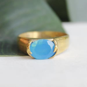May include: A gold-toned ring featuring a large, oval-shaped, light blue gemstone. The gemstone is the focal point, set in a simple, yet elegant, gold band. The ring is displayed on a white surface.
