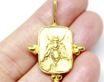 May include: A gold-colored pendant with a rectangular shape and a detailed bee design. The pendant has a loop at the top for hanging and decorative elements on the sides and bottom. The pendant is held in a hand.