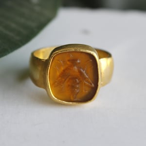 Op de afbeelding: Een gouden ring met een vierkante, amberkleurige steen. De steen is gegraveerd met een gedetailleerde afbeelding van een bij. De ring heeft een dikke band en een gepolijste afwerking, in vintage of antieke stijl.